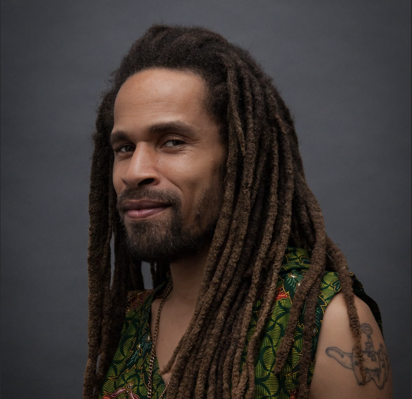 A portrait of a man in semi-profile looking at the camera and smiling while wearing a green patterned sleeveless shirt